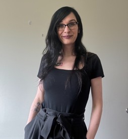 Headshot of Kaelea Shaner, LGBTQ+ Commissioner, smiling in a black shirt and black-framed glasses.