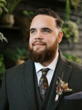 Headshot of Noah Duckett, LGBTQ+ Commissioner, smiling in a black tuxedo.